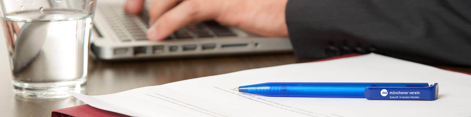 Stift liegt auf Dokument während Person im Hintergrund am Laptop arbeitet